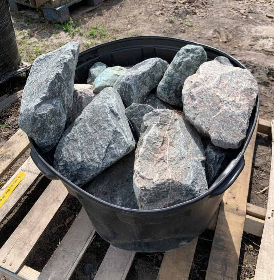 Black bin filled with large gray stones on a wooden pallet outdoors.