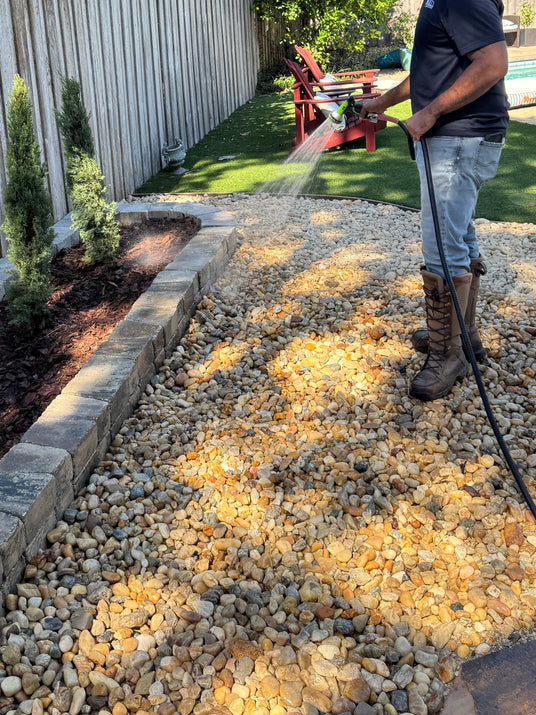 Person using a pressure washer on a gravel area with trees and a wooden fence in the background.