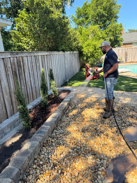 Person using a pressure washer on a gravel area with trees and a wooden fence in the background.